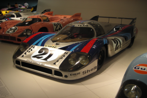 Ein Porsche 917K Le Mans Rennwagen in einem Museum ausgestellt, umgeben von anderen Sportwagen und Menschen, mit einer Wand im Hintergrund und einem Blatt Papier mit Text links daneben.