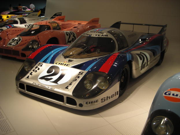 Ein Porsche 917K Le Mans Rennwagen in einem Museum ausgestellt, umgeben von anderen Sportwagen und Menschen, mit einer Wand im Hintergrund und einem Blatt Papier mit Text links daneben.