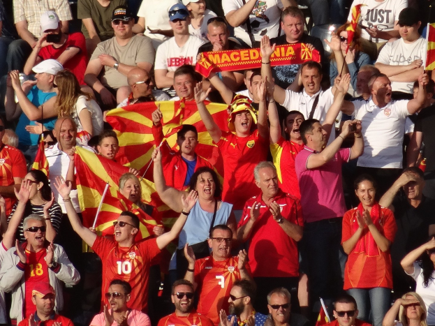 Menge in einem Stadion, die Hände hebt, einige tragen Mützen und Schutzbrillen, halten Fahnen und Schilder mit Text, mit Geländern im Hintergrund.