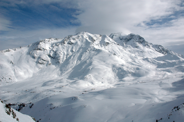 Ein schneebedeckter Berg mit einigen Skifahrern, die hinunterfahren, umgeben von einer bewölkten Himmel und unberührtem Schnee.