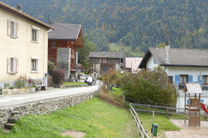 Kleines Dorf mit Häusern, Bäumen, Pflanzen, Gras, einem Holzzaun, einer Rutsche, einem Mülleimer, einer Straße, Fahrzeugen, Polen und einem Hügel im Hintergrund.