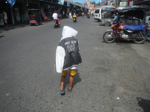 Ein junger Junge geht eine Straße entlang, die von Motorrädern und anderen Fahrzeugen gesäumt ist, flankiert von Gebäuden mit Schildern, unter einem klaren blauen Himmel mit Bäumen und Pfählen im Hintergrund.
