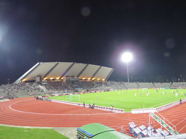 Ein großes Stadion voller Menschen, beleuchtet von Flutlicht, umgeben von Gras, Geländern, Stühlen und anderen Gegenständen, mit dem Himmel im Hintergrund.