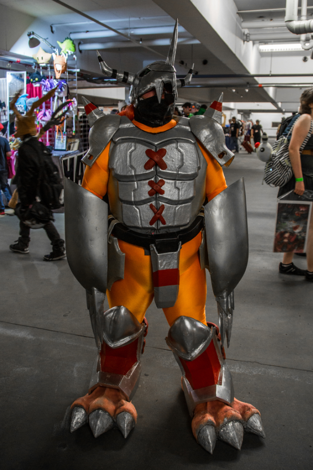 Ein Roboter steht in der Mitte des Bildes umgeben von Menschen, mit einer Wand und Rohren im Hintergrund und Spielzeug auf einem Tisch links.