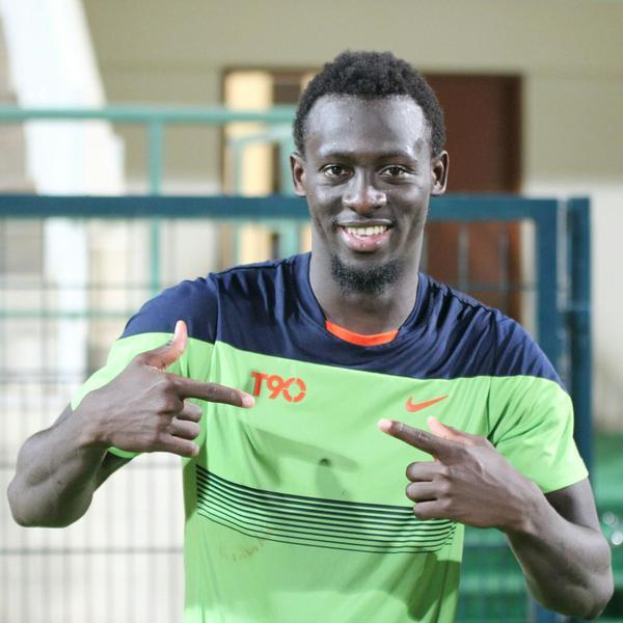 Ein Mann in einem grün-blauen Hemd zeigt mit einem Lächeln in die Kamera, mit einem unscharfen Geländer, einer Wand und einer Tür im Hintergrund.