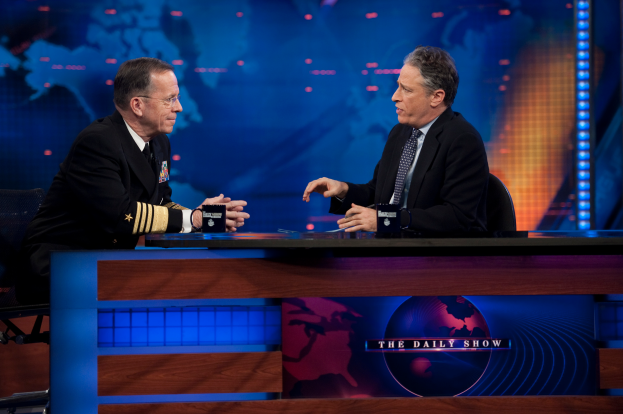 Zwei Männer in schwarzen Blazern, Hemden und Krawatten sitzen an einem Tisch mit Mikrofonen dazwischen und unterhalten sich vor einem Fernsehbildschirm und einem blauen Hintergrund.