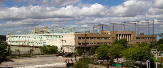 Eine Straße mit Fahrzeugen, gesäumt von Bäumen und Straßenlaternen, führt zu einem großen Gebäude in der Nähe eines Gewässers.