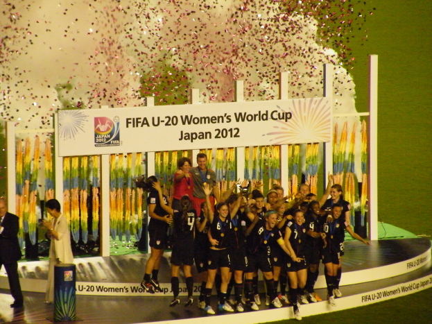 Eine Gruppe von Frauenfußballspielerinnen in schwarzen Trikots feiert ihren Sieg auf einer Bühne, umgeben von einer Menge mit Kameras, mit einem dekorierten Hintergrund und einem grasbewachsenen Boden.