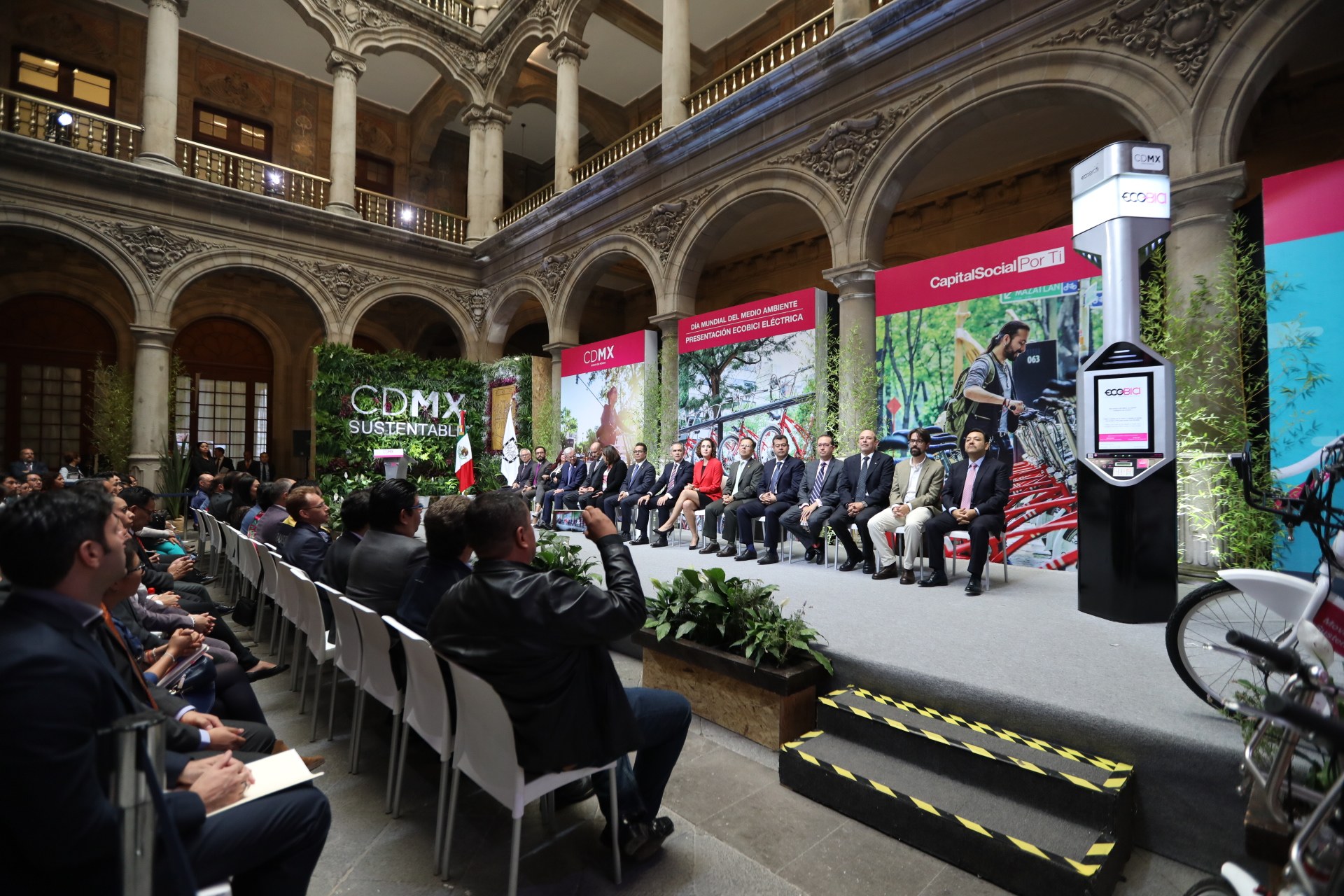 Eine Gruppe von Menschen sitzt in Stühlen in einem Gebäude, mit einem Mann auf einer Bühne davor. Rechts im Bild stehen Fahrräder, und im Hintergrund gibt es Werbetafeln, Pflanzen, Säulen, Geländer, Lampen, Fenster und eine Wand.