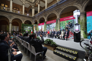 Eine Gruppe von Menschen sitzt in Stühlen in einem Gebäude, mit einem Mann auf einer Bühne davor. Rechts im Bild stehen Fahrräder, und im Hintergrund gibt es Werbetafeln, Pflanzen, Säulen, Geländer, Lampen, Fenster und eine Wand.