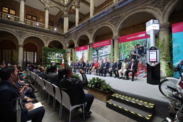 Eine Gruppe von Menschen sitzt in Stühlen in einem Gebäude, mit einem Mann auf einer Bühne davor. Rechts im Bild stehen Fahrräder, und im Hintergrund gibt es Werbetafeln, Pflanzen, Säulen, Geländer, Lampen, Fenster und eine Wand.