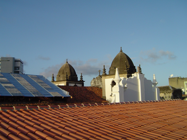 Stadtansicht mit Gebäuden im Vordergrund, einem blauen Himmel im Hintergrund und Solarpanelen auf einem Dach.