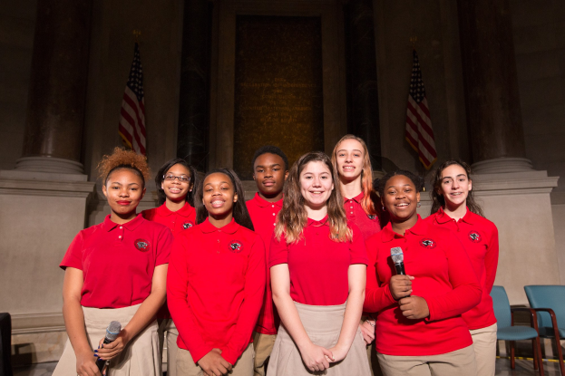 Gruppe junger Frauen in roten Shirts, die strahlend Mikrofone halten und vor Stühlen, Säulen, Flaggen und einer Wand stehen, wahrscheinlich Teil eines nationalen Jugendchors.