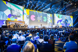 Eine große Menschenmenge sitzt auf Stühlen vor einer Bühne auf der Deutschen Internet-Konferenz 2019, mit einem Tisch voller Gegenstände links und Bildschirmen und Lichtern im Hintergrund.