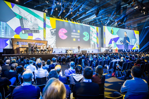Eine große Menschenmenge sitzt auf Stühlen vor einer Bühne auf der Deutschen Internet-Konferenz 2019, mit einem Tisch voller Gegenstände links und Bildschirmen und Lichtern im Hintergrund.