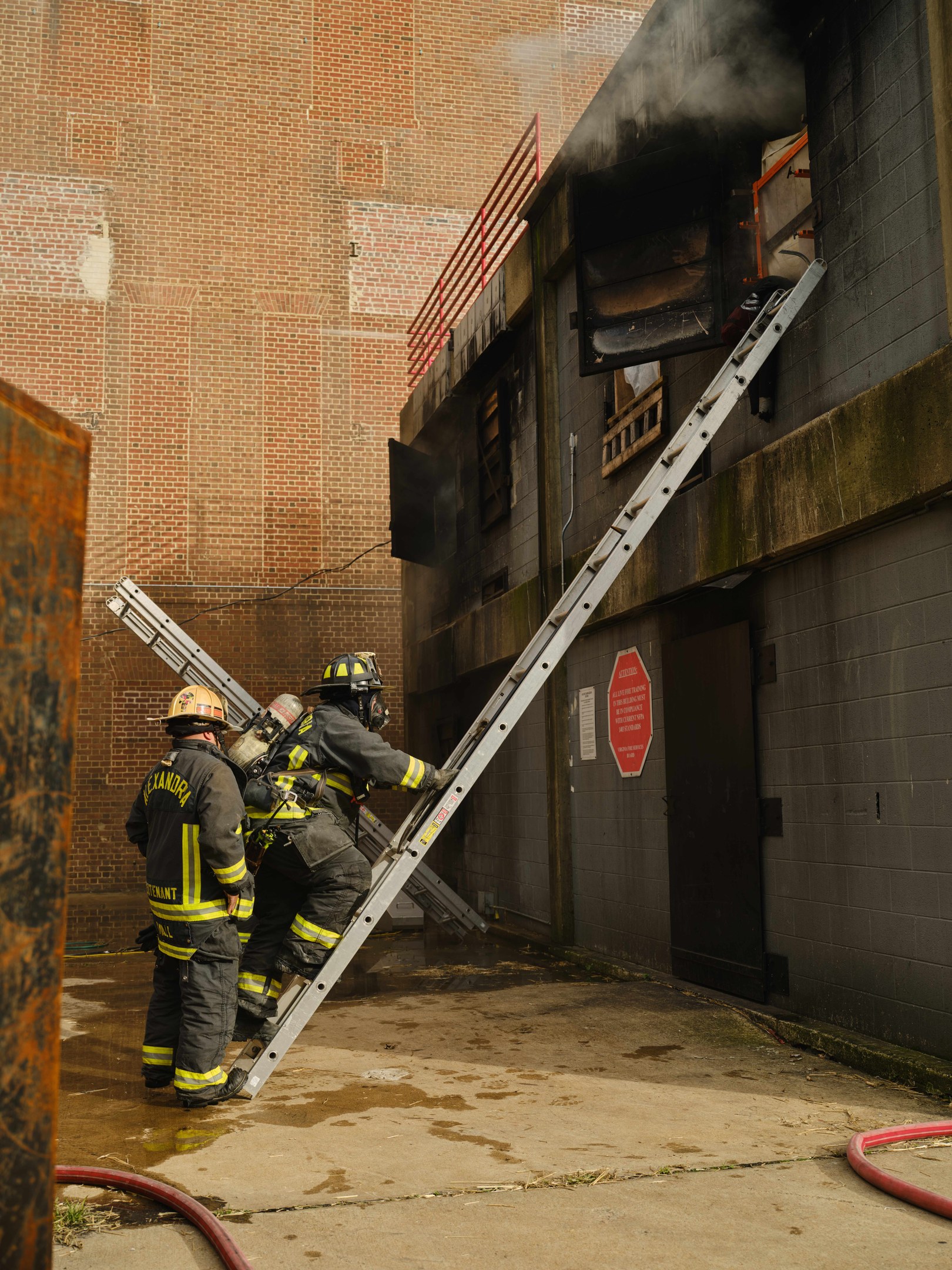 Feuerwehrleute in Helmen, die eine Leiter zu einem raucherfüllten Gebäude hinaufsteigen, mit Rohren und einem Metallobjekt in der Nähe.