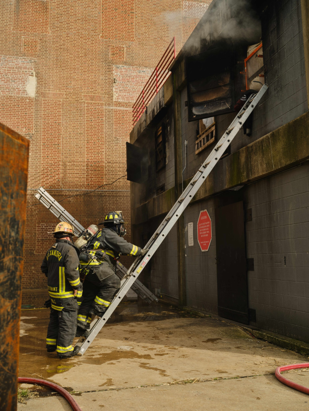 Feuerwehrleute in Helmen, die eine Leiter zu einem raucherfüllten Gebäude hinaufsteigen, mit Rohren und einem Metallobjekt in der Nähe.