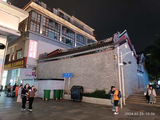 Menschen gehen nachts eine Straße entlang, beleuchtet von Straßenlaternen und umgeben von Gebäuden mit Fenstern, Schildern, Mülltonnen, Pflanzen und Bäumen unter einem bewölkten Himmel, einige tragen Taschen.
