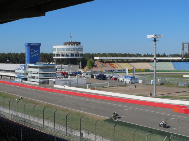 Eine Gruppe von Menschen auf Motorrädern auf einer Rennstrecke, umgeben von einem Zaun, Fahrzeugen, einem Gebäude, Schildern, Pfosten, einem Stadion, Lampen, Bäumen und einer bewölkten Himmel.