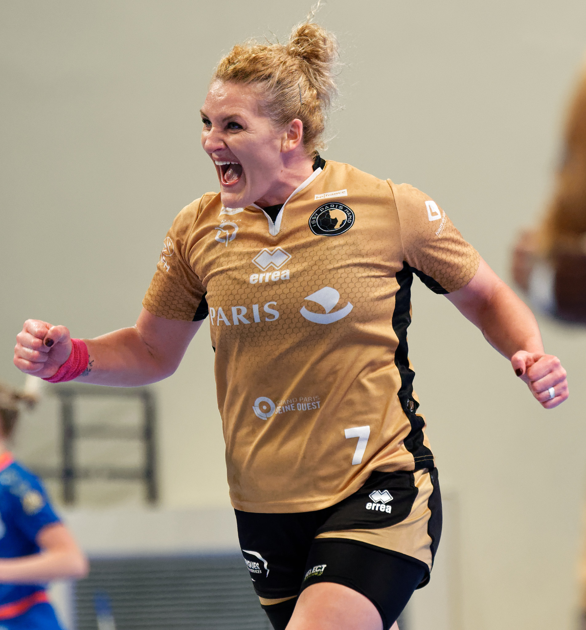 Eine Frau in braunem T-Shirt und schwarzen Shorts rennt auf einem Futsalplatz, feiert mit triumphaler Miene, während zwei andere Frauen im Hintergrund unscharf sind.