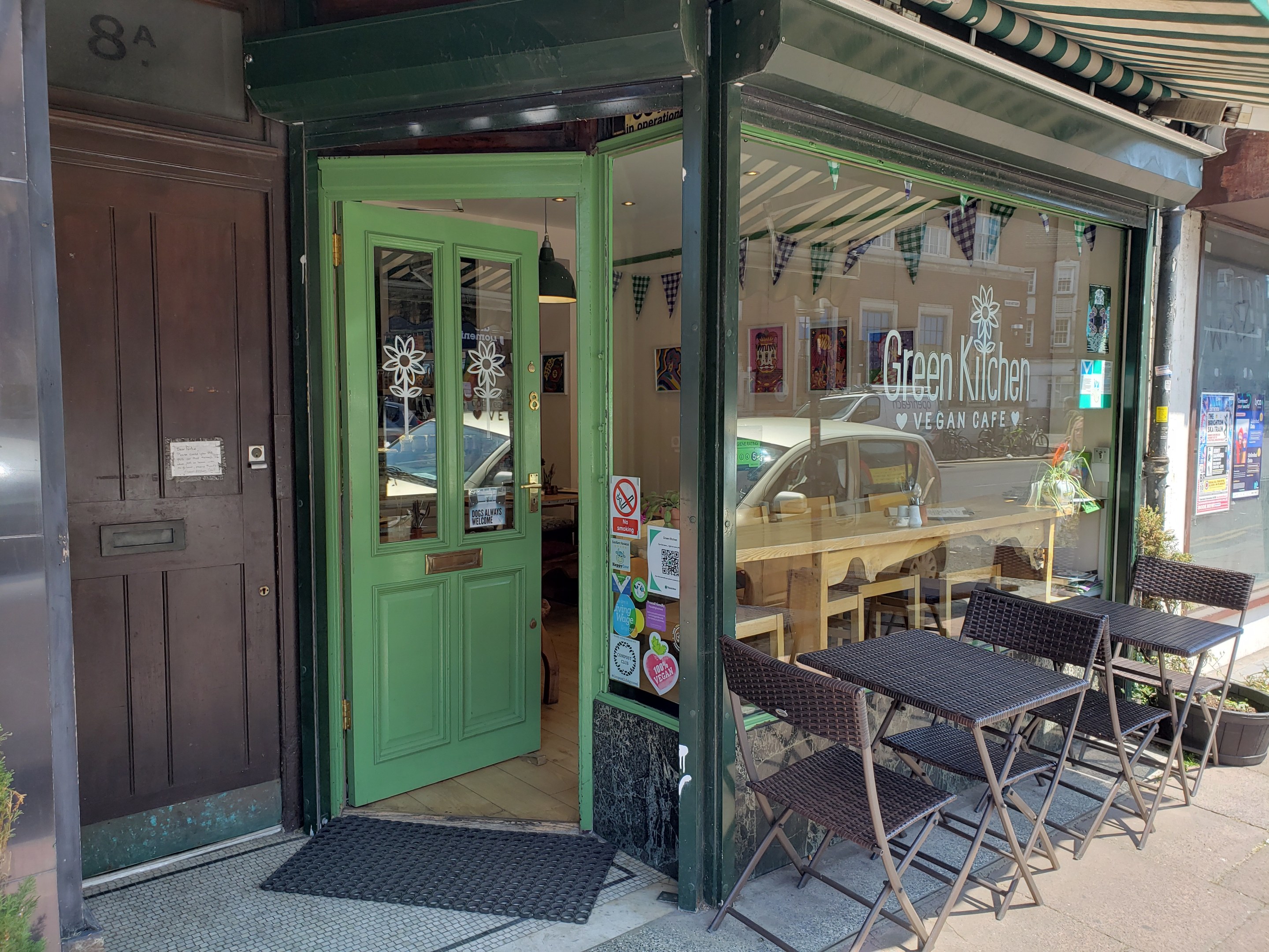 Außensitzbereich des Green Kitchen Vegan Cafe mit Tischen, Stühlen, Pflanzen, einer Fußmatte und einer Glastür mit Postern; drinnen sind Tische, Stühle und Deckenleuchten zu sehen.