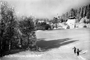 Zwei Menschen Langlaufski fahren im Schnee, umgeben von Bäumen und Häusern im Hintergrund, schwarz-weißes Bild mit Text unten.