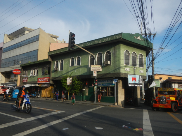 Eine belebte Stadtstraße mit einem grünen Gebäude im Hintergrund, Fahrzeugen, Fußgängern, einer Ampel, bewölktem Himmel und Oberleitungen.