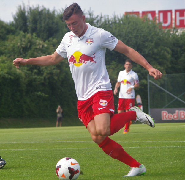 Ein Mann in einem Fußballtrikot kickt einen Ball auf einem grünen Feld mit Bäumen und einem klaren blauen Himmel im Hintergrund, mit einem Schild mit der Aufschrift «RB Leipzig» sichtbar.