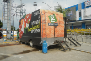 Ein großer Lastwagen parkt vor einem Gebäude mit einer Tafel, auf der "Teilen Sie eine positiv überladene Erfahrung" steht, mit Stufen, einem Zylinder auf dem Boden, Geländern, Strom- und Lichtmasten, Bäumen und einem bewölkten Himmel.