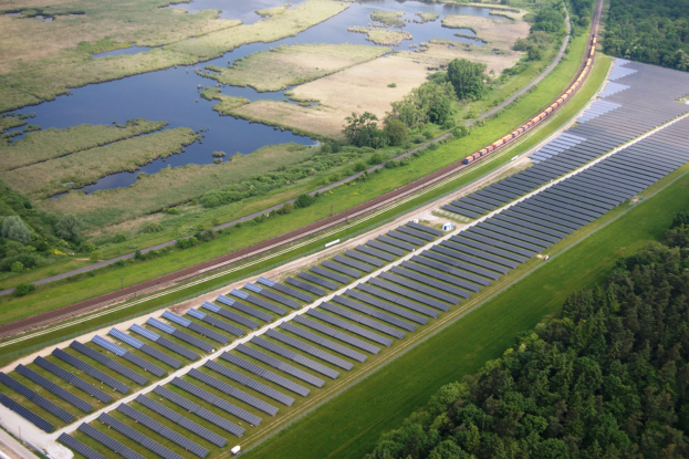 Luftaufnahme einer Solar-Farm mit Panelen in einem Feld, umgeben von Bäumen, Gras, Wasser und einem Zug auf einer Bahnstrecke.