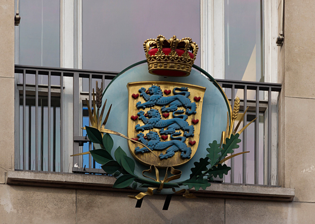 Wappen der britischen Königsfamilie auf der Fassade eines Gebäudes dargestellt, begleitet von einem Kronen- und Blätter-Logo, mit Fenstern und Metallgittern im Hintergrund.