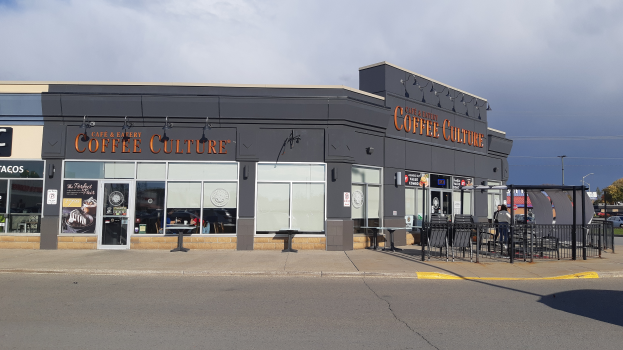 Ein Kaffeehaus an einer Straßenecke mit einer Straße im Vordergrund, einem Gebäude im Hintergrund, einigen Fahrzeugen, Bäumen und einem blauen und weißen Himmel.