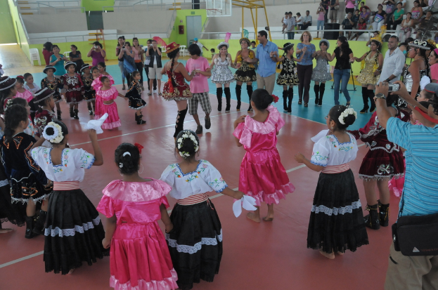 Eine Gruppe von Kindern in Kostümen tanzt in der Mitte des Bildes, während das Publikum von der oberen rechten Ecke zusieht.