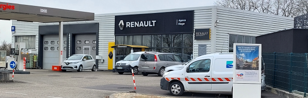 Ein Renault-Autohaus mit parkenden Autos vorne, umgeben von einem Metallzaun, einem Schild, Pfosten, einer Texttafel, Bäumen und einem bewölkten Himmel.