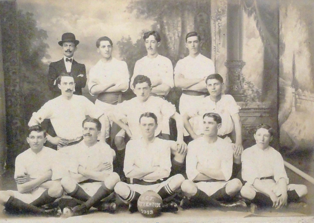 Ein Schwarz-Weiß-Foto einer Fußballmannschaft, mit einigen Spielern, die sitzen und einige, die stehen, einem Ball in der Mitte und Bäumen und einem klaren Himmel im Hintergrund, mit Text unten.