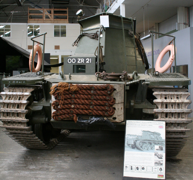 Ein Panzer steht auf dem Boden in einem Raum mit einer Tafel, mehreren Bildern, einer Wand, Säulen, Lampen, einem Schuppen und Stangen im Hintergrund.