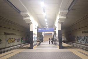 Eine U-Bahn-Station mit Graffiti an den Wänden, ein Zug auf den Schienen, Menschen auf dem Bahnsteig, Bänke, Pfeiler, ein Namensschild, Deckenleuchten und Treppen im Hintergrund.