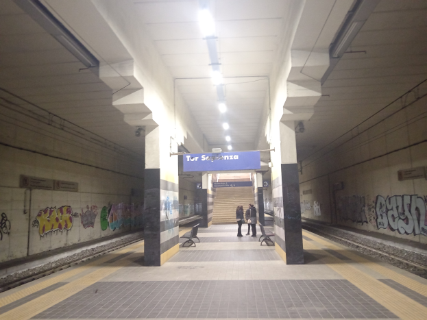 Eine U-Bahn-Station mit Graffiti an den Wänden, ein Zug auf den Schienen, Menschen auf dem Bahnsteig, Bänke, Pfeiler, ein Namensschild, Deckenleuchten und Treppen im Hintergrund.
