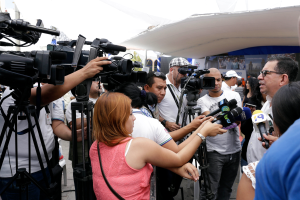 Eine Gruppe von Menschen umringt Reporter mit Mikrofonen und Kameras, mit einem Zelt und Bannern im Hintergrund unter einem sichtbaren Himmel.