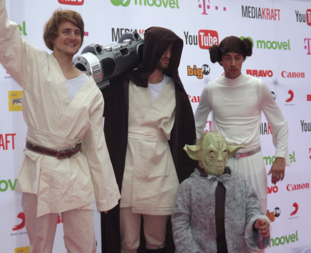 Gruppe von Menschen in Star-Wars-Kostümen auf einem roten Teppich posend, wobei eine Person ein Objekt und ein Banner mit Text und Logos im Hintergrund hält.