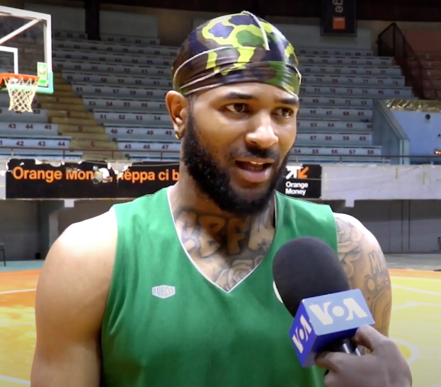 Ein Mann in einem grünen Tanktop hält ein Mikrofon und steht auf einem Basketballfeld mit einem Netz auf der linken Seite und Treppen mit Geländern im Hintergrund, wahrscheinlich spricht er mit den Medien über die bevorstehende Basketballsaison.