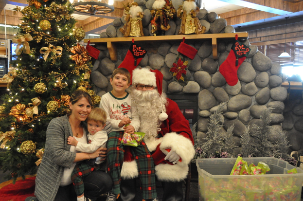 Eine Frau, Kinder und ein Mann als Santa Claus mit Brille und Handschuhen stehen in der Nähe eines geschmückten Weihnachtsbaums, einer Wand mit Steinen, Spielzeugen, Dekorationen und einem Korb mit Gegenständen, mit Lichtern im Hintergrund.