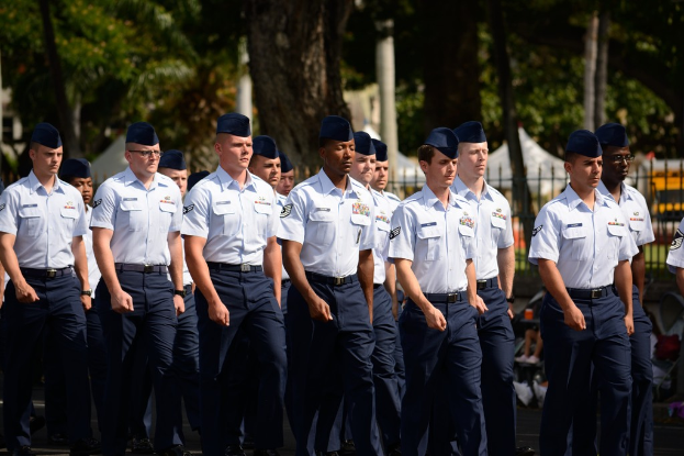 Männer in Uniform auf einer Straße mit einem Eisengeländer dahinter, Bäume und Masten im Hintergrund.