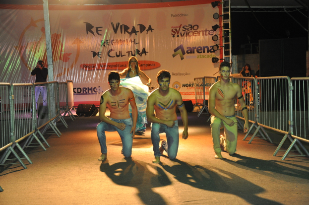 Drei Jungen ohne Hemd knien auf dem Boden während eines kulturellen Ereignisses, mit Grills auf beiden Seiten und einem großen Banner im Hintergrund, während jemand auf der linken Seite ein Foto macht.