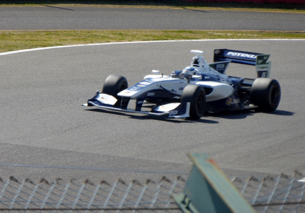 Ein Mann fährt ein Rennauto auf einer Rennstrecke mit einem Metallzaun am Boden und Gras auf dem Boden, mit einer texttragenden Wand im Hintergrund.