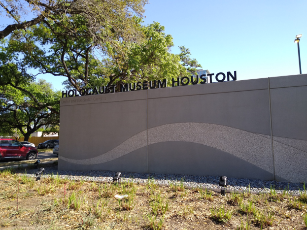 Außenansicht des Houstoner Holocaust-Museums mit eingravierter Textwand, Gedenksteine, Landschaftsgestaltung, vorbeifahrenden Fahrzeugen und einem bewölkten Himmel.