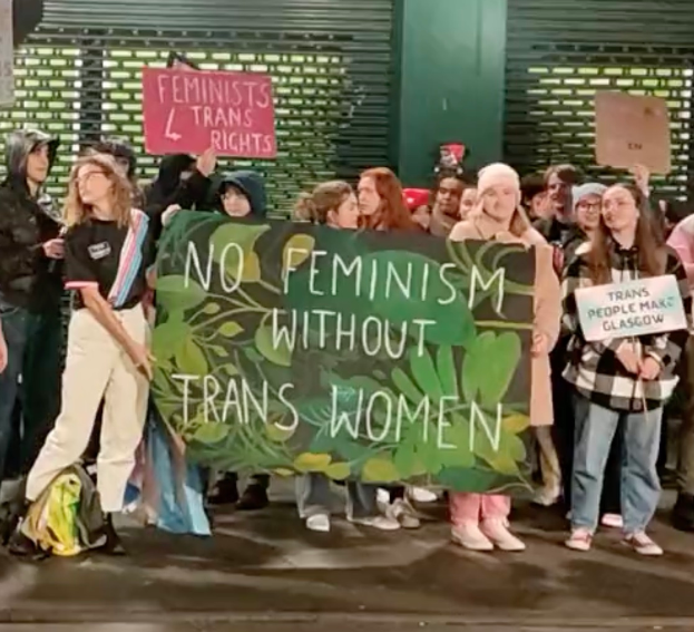 Eine Gruppe von Menschen hält ein Transparent mit der Aufschrift "Kein Feminismus ohne Trans-Frauen" und verschiedene Schilder in der Hand und steht an einem öffentlichen Ort mit einer Wand und einem Rollladen im Hintergrund.