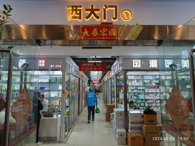 Innenansicht einer chinesischen Apotheke mit Glaswänden, Regalen voller Artikel, verstreuten Kartons, einer Pflanze links und einer Namensschild oben.
