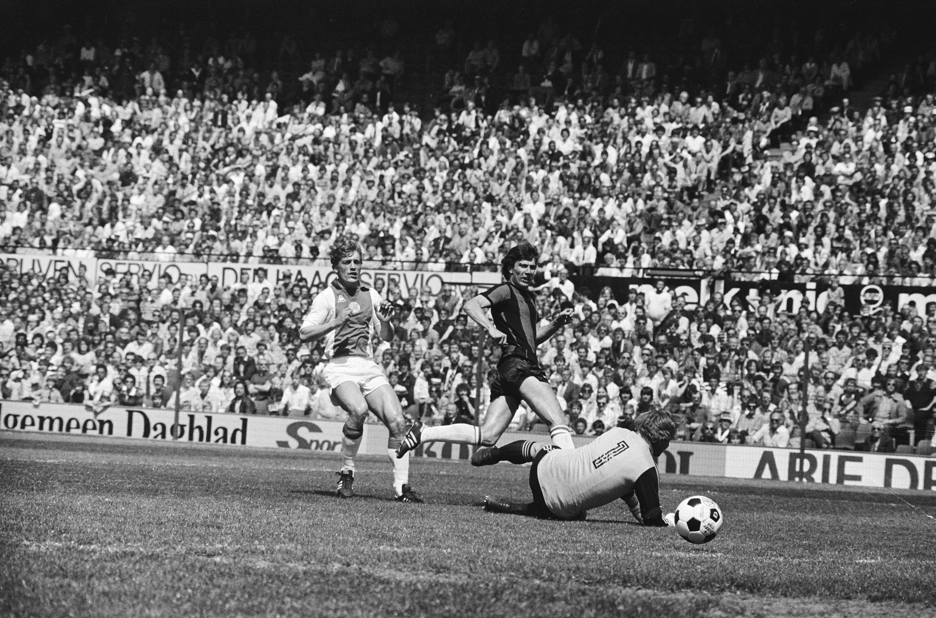 Schwarzes und weißes Foto von Fußballspielern in Aktion auf einem Feld mit Zuschauern im Hintergrund und einem Bundesliga-Banner 1978-79 Ajax vs. Hannover 96.