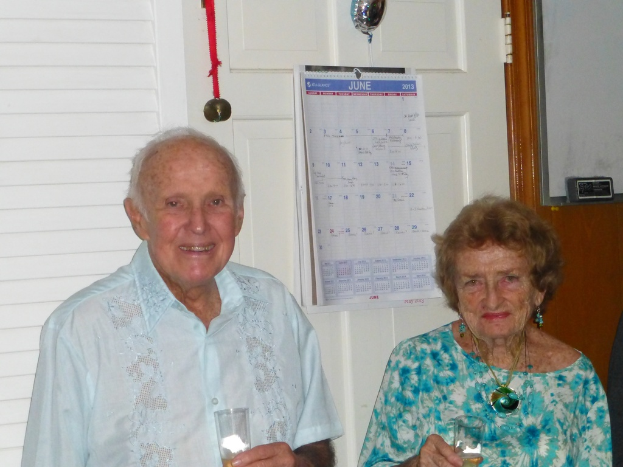 Ein älterer Mann in einem Hemd hält ein Glas und lächelt, neben einer Frau in einem blauen Kleid mit Ohrringen und einer Kette stehend, ebenfalls ein Glas haltend, mit Medaillen und einem Kalender auf der weißen Wand im Hintergrund.
